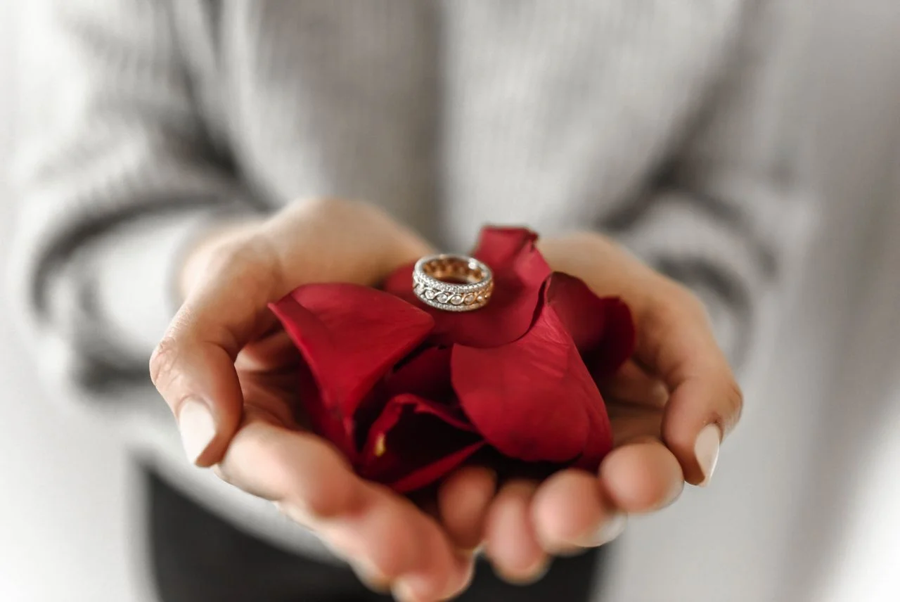 Diamond ring resting on red rose petals held in hands.